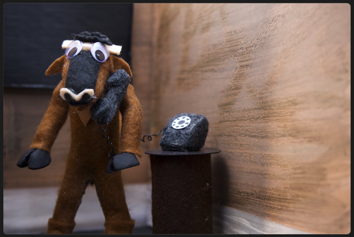 A felt horned character holding a handmade rotary telephone receiver, standing beside the telephone on a rusted pedestal.