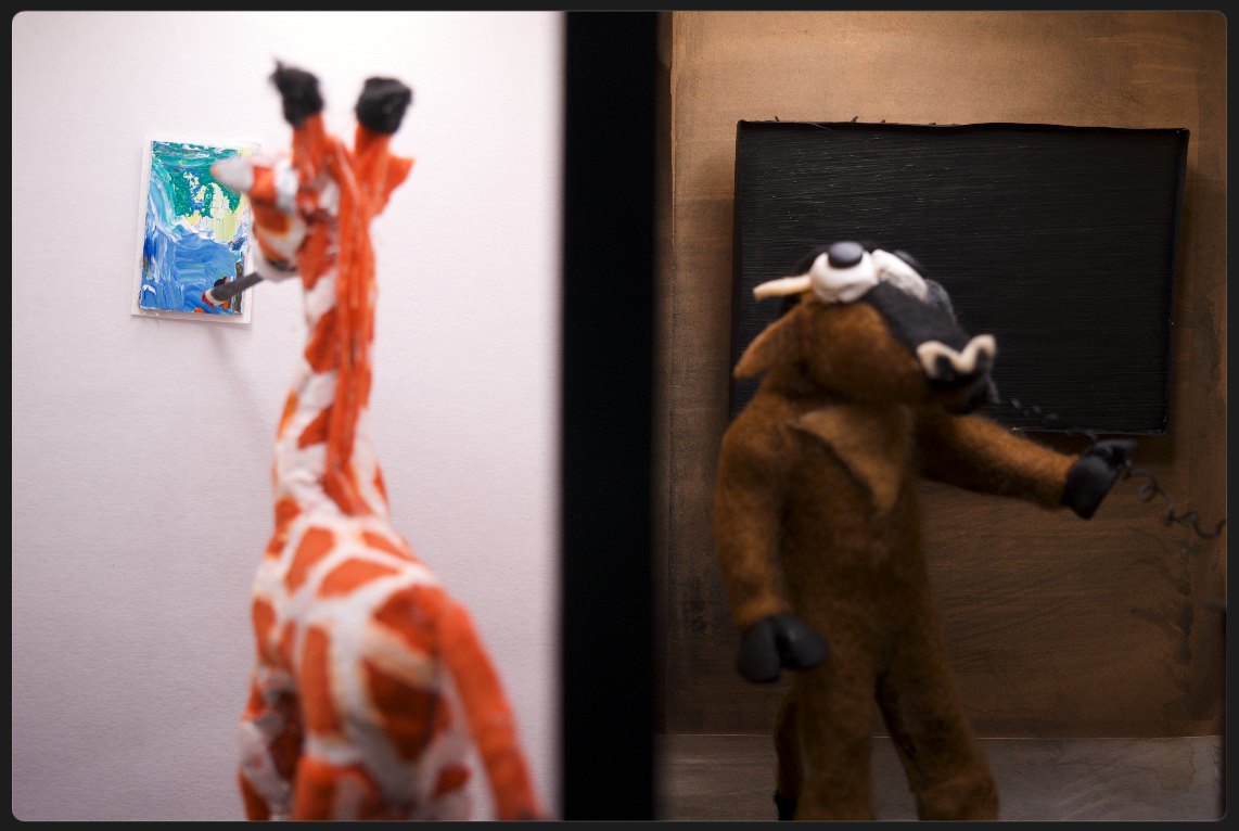 Two handmade felt characters in a built interior: a giraffe looking at a small painting hung on a white wall, and a horned figure writing at a blackboard.