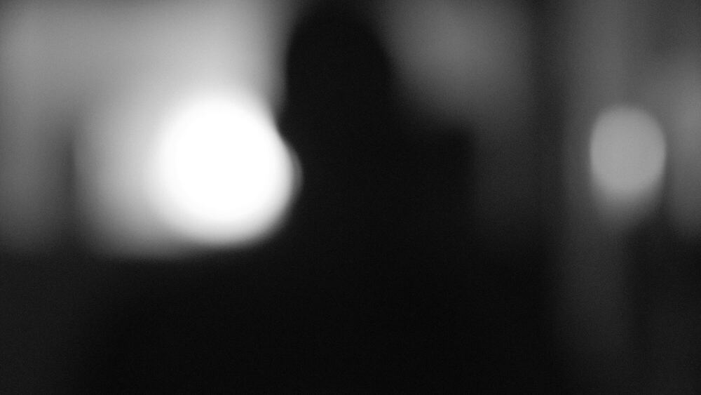 A grey, almost abstract frame in which a dark human shape and two soft discs of light are diffused past recognition by textured glass.