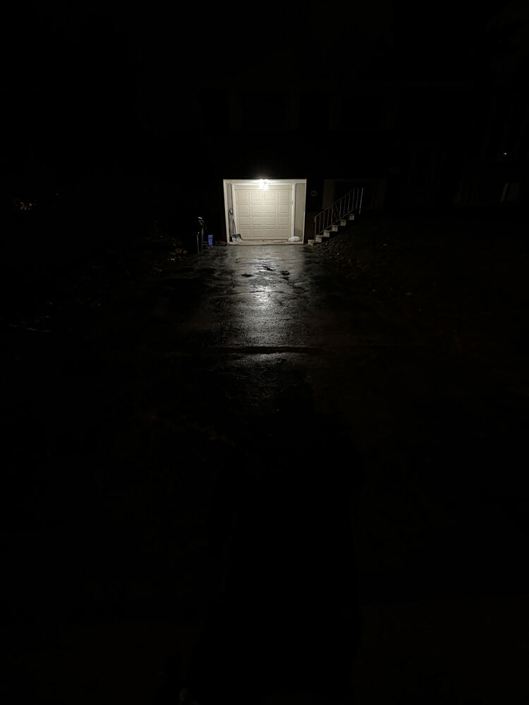 A nearly black vertical frame with a small bright doorway far away at the top, its light laid out along a wet drive leading toward the camera.