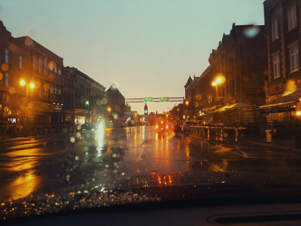 A small-town main street at dusk seen from inside a car, rain beaded on the windshield, storefront and street lights doubled in the wet road and a teal sky above the brick.