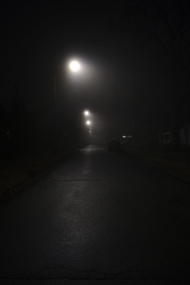 A dark residential street photographed down its centre line at night, with one bright streetlight overhead and two smaller ones receding into the distance.