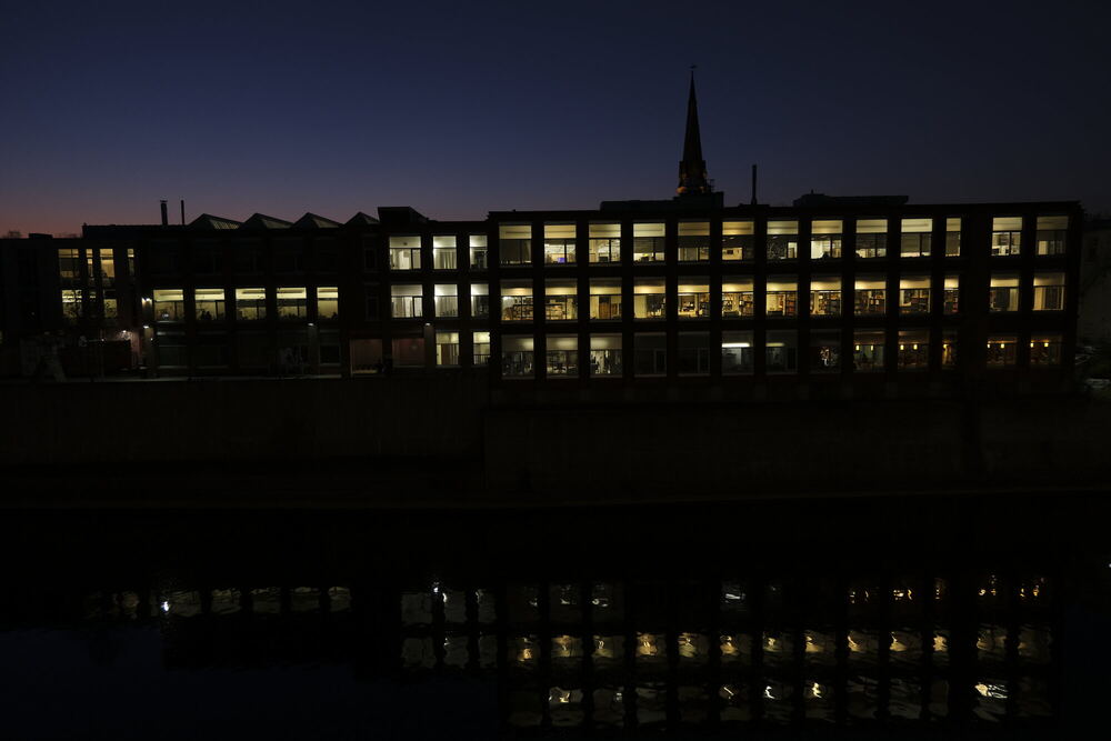 A long three-storey building at blue hour, every window lit from inside, standing above a dark river that breaks the grid of windows into a looser pattern of light.
