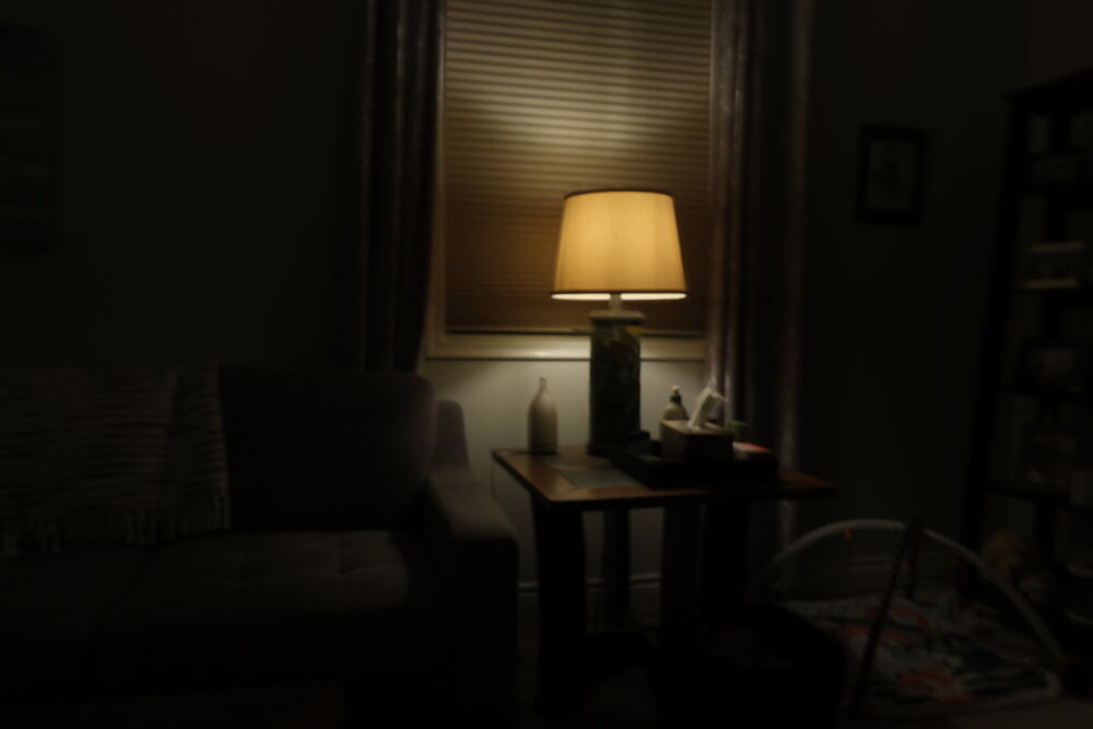 A living room lit by one table lamp beside a blinded window, photographed from an adjoining unlit room so that the foreground falls away into black.