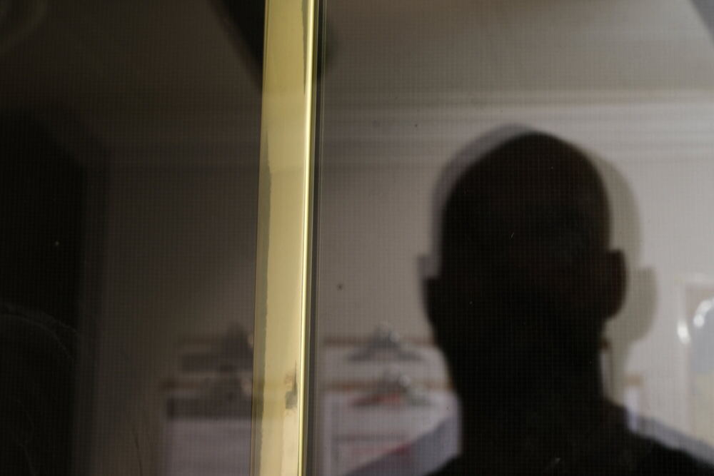 The soft dark silhouette of a head and shoulders pressed against a white screen door, the mesh rendering the figure as a flat grey shape with no features.