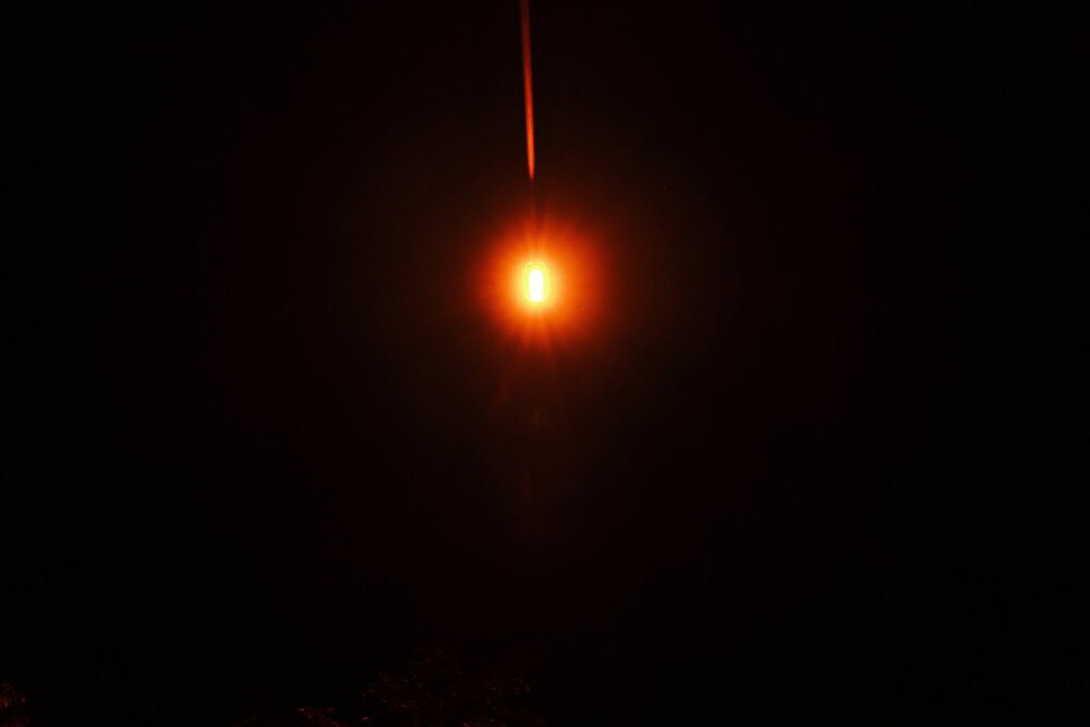 A single small point of warm orange light burning in an otherwise entirely black frame, with a faint vertical flare rising from it.