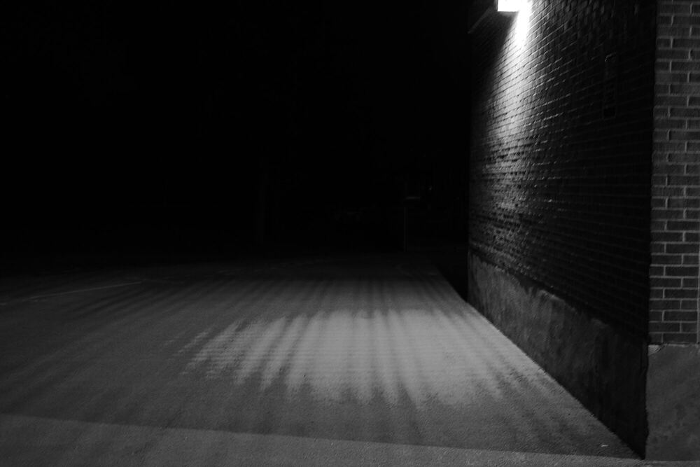 A narrow alley at night in near monochrome, a single wall-mounted lamp high on the right throwing a long striped wash of light down the paving.