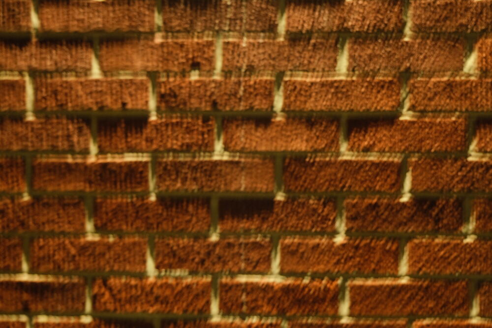 A close view of an orange brick wall lit from directly above, so that every course of mortar casts a hard shadow and the texture of each brick reads as relief.