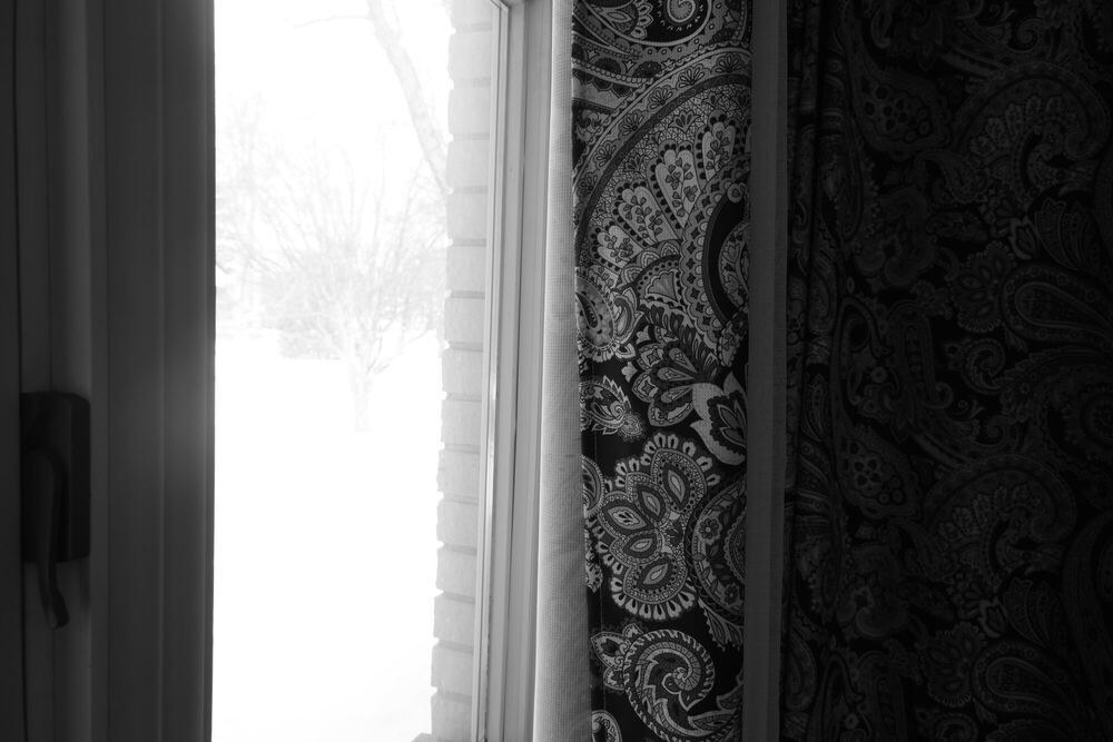 Black and white: a vertical slot of blown-out winter daylight beside a window frame, with a heavily patterned paisley curtain filling the right half of the frame in deep black.